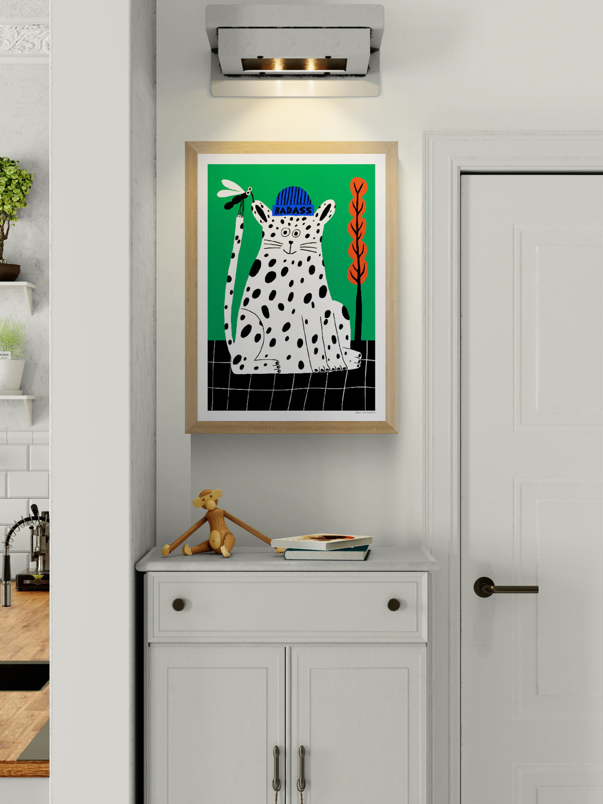 Badass by Dans les Dents, vertical giclée art print of a playful black-spotted white cat wearing a blue beanie, holding a dragonfly against a bright green background with orange plants. Bold, contemporary wall art.