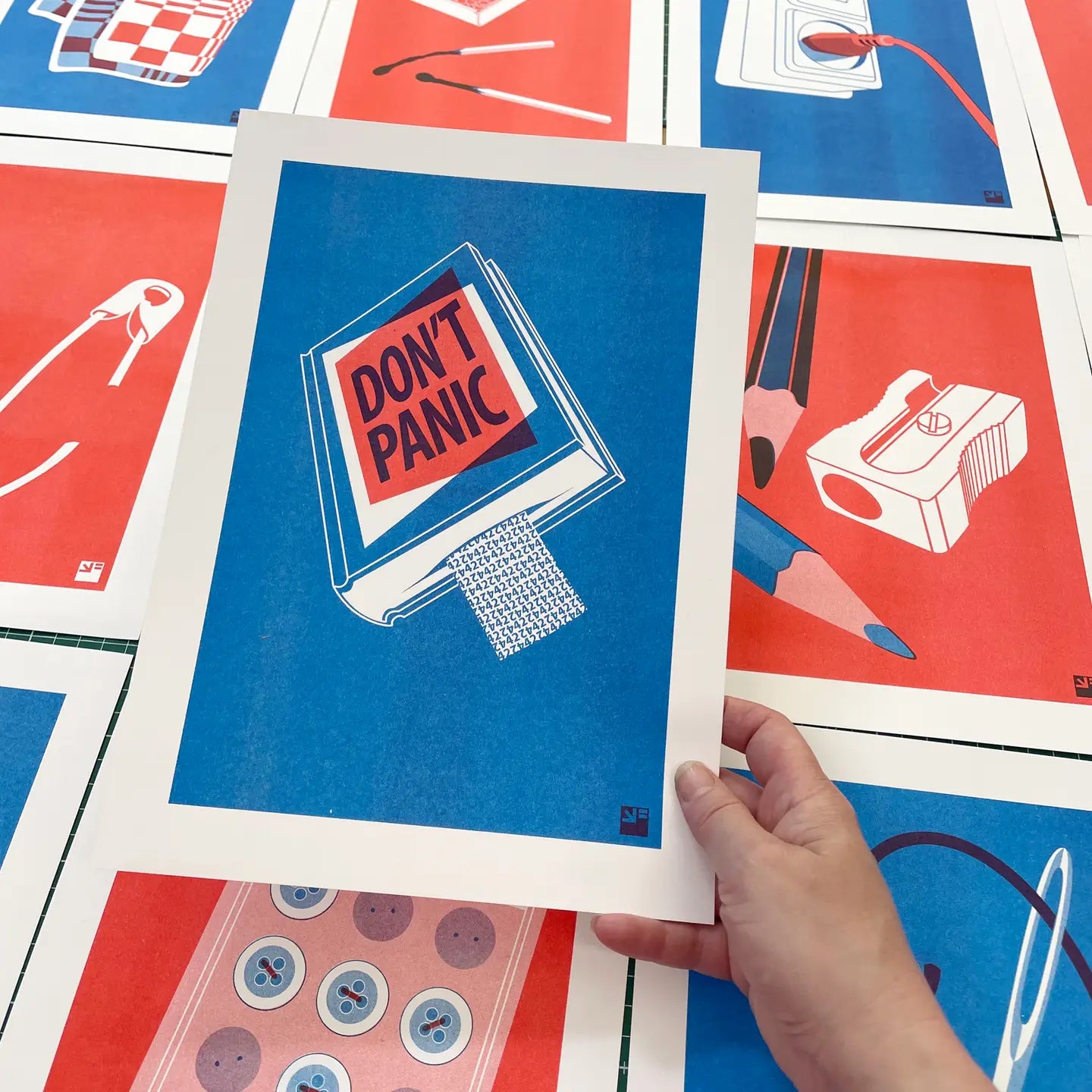 Risograph art print titled Book by VrijFormaat, featuring a blue book cover with bold red and black lettering reading “DON’T PANIC”, inspired by The Hitchhiker’s Guide to the Galaxy. Minimalist design printed in red and blue vegetable-based inks on Biotop paper, A4 size.
