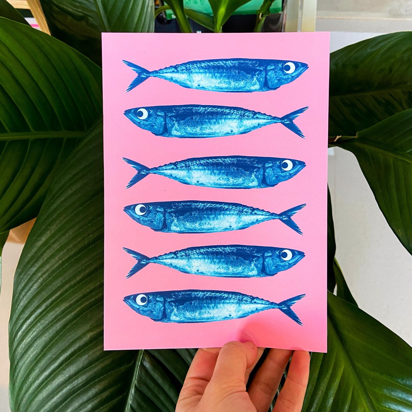 Bright pink and blue offset print of sardines by Dutch illustrator PTRVRKD – playful modern seafood illustration with seven blue fish on bold pink background, printed with eco-friendly soy inks on 250 gsm Biotop paper, handmade in the Netherlands – quirky small art print for kitchens or walls.