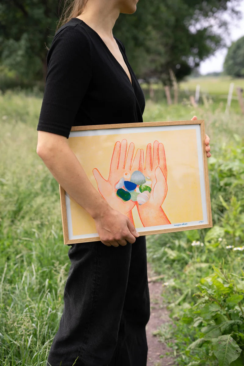 Coquillages by French illustrator Margaux Othats, A3 (29.7 x 42 cm) risograph print in delicate pastel colors. The artwork shows two hands holding seashells and sea glass, evoking summer memories and tenderness. Printed on 150g Munken Print White paper, limited edition of 150 signed and numbered copies, made in Belgium, unframed, packaged with protective sleeve and cardboard backing.