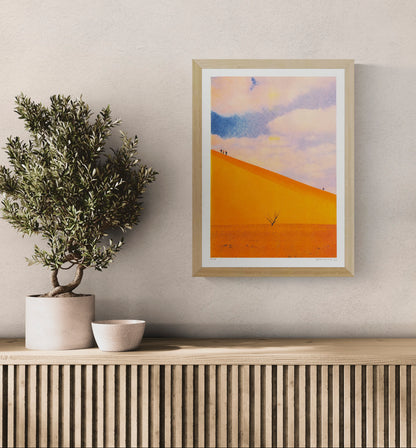 A3 portrait risograph by PTRVRKD showing vivid orange desert dunes, tiny walking figures, a lone tree and a soft blue-pink sky, ideal for modern wall art and home decor.