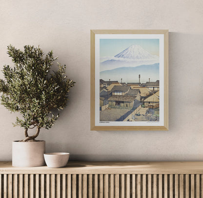 Morning in Motoyoshiwara by Kawase Hasui, Japanese woodblock-style landscape featuring snow-covered Mount Fuji above traditional village rooftops in soft blue and beige tones. Vertical digital art print for serene wall decor.