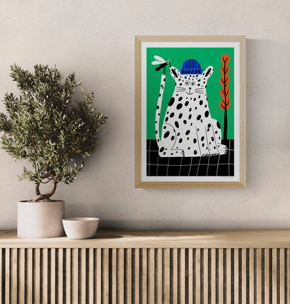 Badass by Dans les Dents, vertical giclée art print of a playful black-spotted white cat wearing a blue beanie, holding a dragonfly against a bright green background with orange plants. Bold, contemporary wall art.