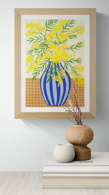 Botanical digital art print titled “Mimosa” by Belgian illustrator Leens, featuring vibrant yellow mimosa flowers in a blue striped vase on a tiled table, printed on textured fine art paper in A4 size.