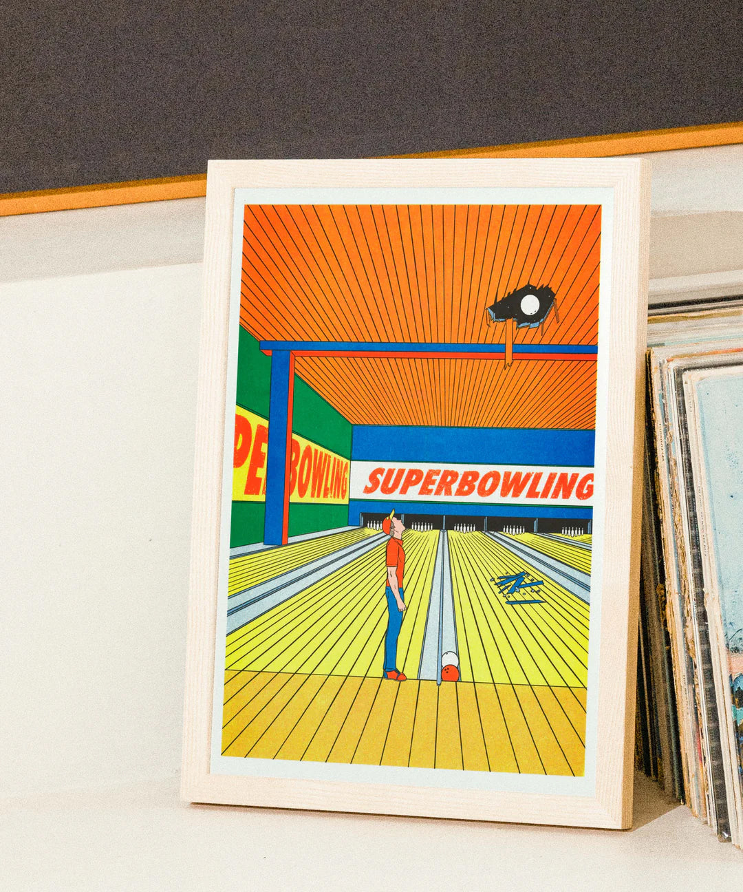 Risograph art print titled Super Bowling by French illustrator Simon Bailly, showing a man in a bowling alley looking up as a bowling ball crashes through the ceiling. The scene is drawn in a bold comic style with vivid orange, blue, and yellow colors, capturing humor and everyday absurdity in a clean, graphic composition.