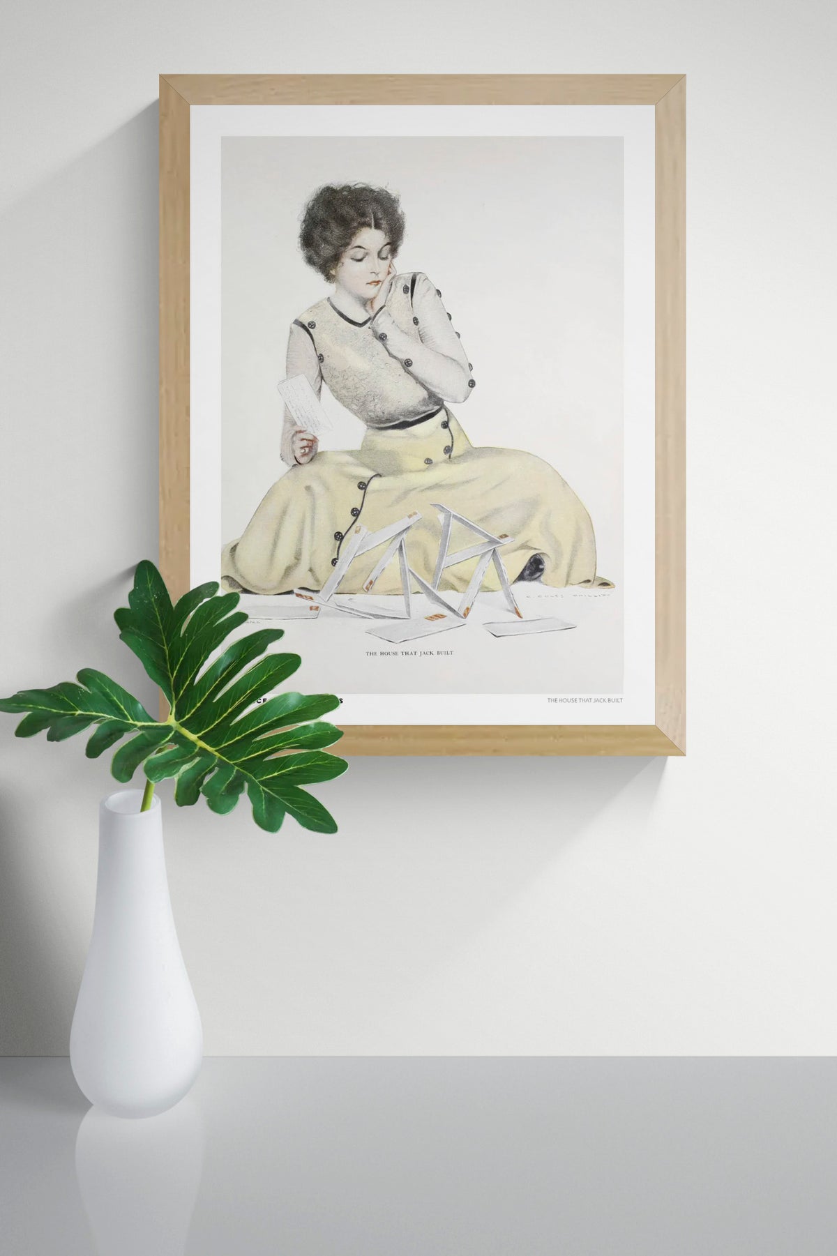 Vintage illustration by Clarence Coles Phillips showing a seated woman in a pale yellow dress holding a letter beside a collapsed house of cards. Soft pastel colors, Edwardian style, vertical digital wall art print.